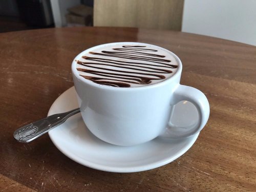 A white cup of hot chocolate with chocolate drizzle on top, sitting on a saucer with a spoon beside it on a wooden table.