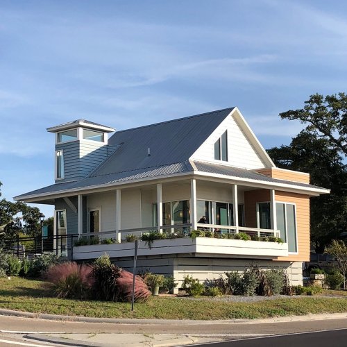 A modern two-story house with a metal roof, large windows, and a wraparound porch, surrounded by greenery.