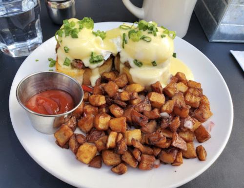 A plate of eggs Benedict topped with hollandaise sauce and green onions, served with crispy diced potatoes and ketchup.