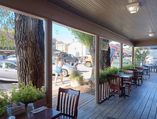 Outdoor dining area with wooden tables, large trees, and a view of a busy street with pedestrians and parked cars.