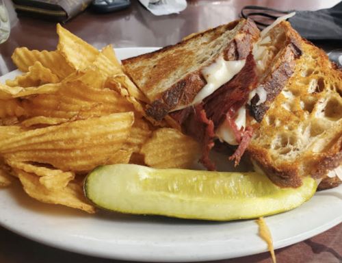 A sandwich with layers of meat and cheese on toasted bread, served with potato chips and a pickle on the side.
