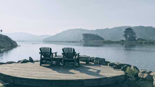 Two Adirondack chairs on a wooden deck by a misty river, surrounded by hills and trees in the background.