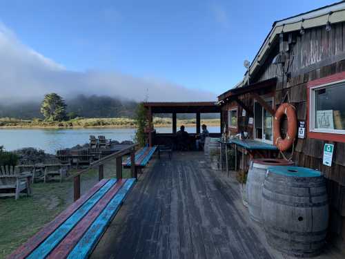 A rustic wooden building by a calm river, with outdoor seating and fog rolling over the hills in the background.