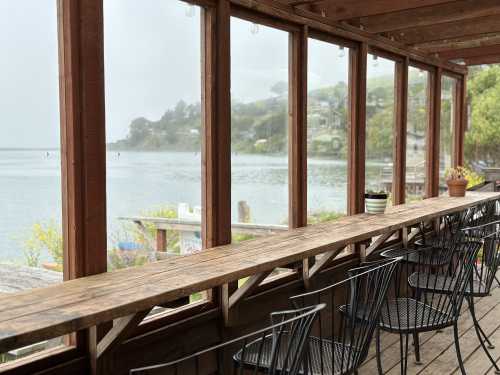 A wooden bar with black chairs overlooks a foggy waterfront and distant houses through large windows.