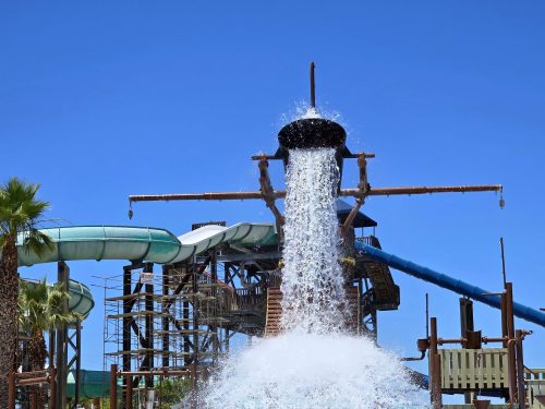 A water slide structure with a large bucket pouring water, surrounded by palm trees and a clear blue sky.