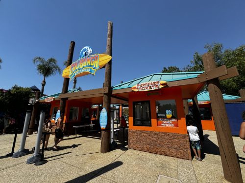 Ticket booth at a water park with a colorful sign, surrounded by palm trees and clear blue skies.