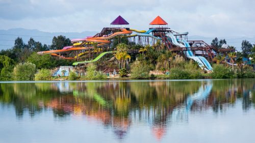 Colorful water slides tower over a lake, reflecting vibrant hues in the water, surrounded by lush greenery.