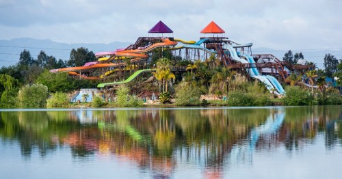 A colorful water park with multiple slides reflected in a calm lake, surrounded by greenery and mountains in the background.