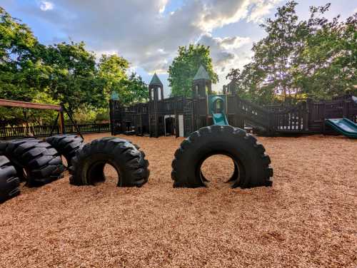 Playground with a wooden structure, green slides, and large tire obstacles on a wood chip surface under a cloudy sky.