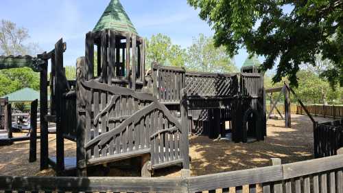 A wooden playground structure with towers and slides, surrounded by trees and a gravel surface.