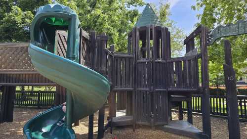 A playground structure featuring a green slide and wooden fort-like design, surrounded by trees and blue sky.