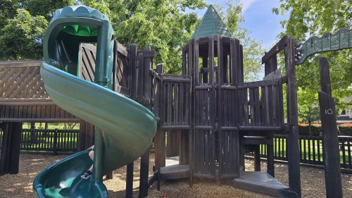 A playground structure featuring a green slide and wooden fort-like design, surrounded by trees and blue sky.