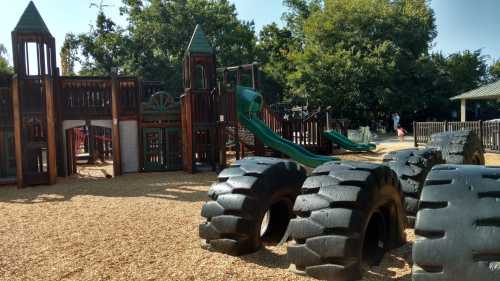 A playground with wooden structures, slides, and large tires on a gravel surface, surrounded by trees.