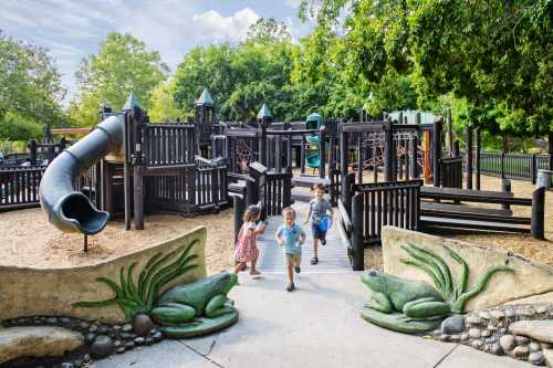 Children play on a wooden playground with slides and climbing structures, surrounded by greenery and frog sculptures.