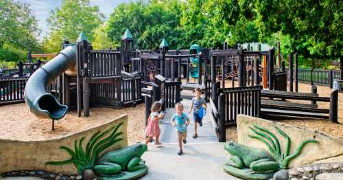 Children play and run in a vibrant playground with wooden structures and a slide, surrounded by greenery.