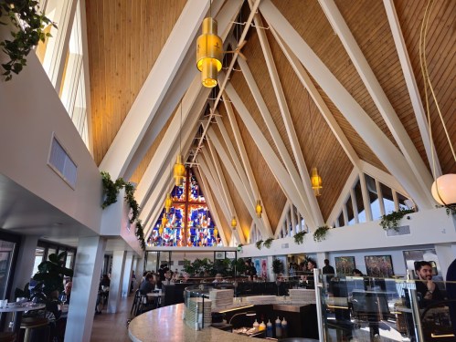 Interior of a modern restaurant featuring a high wooden ceiling, large windows, and colorful stained glass.
