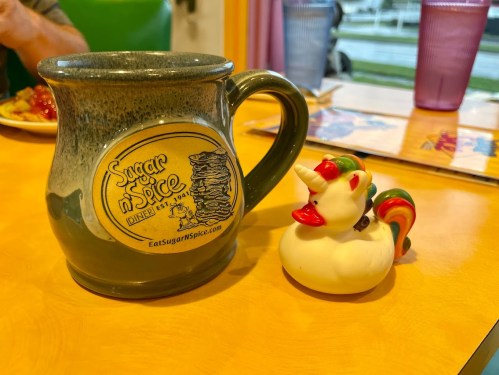 A green mug with a logo next to a colorful rubber duck with a unicorn horn on a bright yellow table.