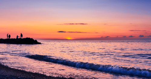 Silhouettes of people on a rocky shore at sunset, with calm waves and a colorful sky reflecting on the water.