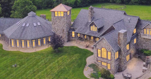 Aerial view of a large stone house with multiple sections, surrounded by green grass and trees, illuminated windows at dusk.