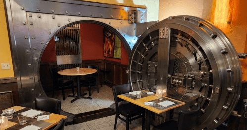 A restaurant interior featuring a large, metallic vault door and a cozy dining area with tables and chairs.