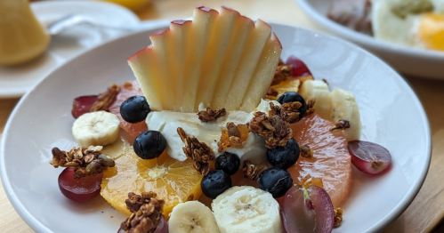 A colorful fruit bowl featuring sliced apples, bananas, grapes, blueberries, and granola on a bed of citrus slices.