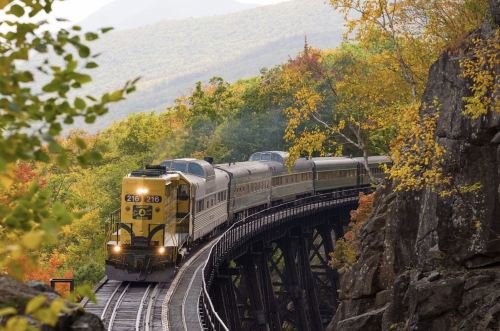 A train travels along a curved track surrounded by vibrant autumn foliage and rocky terrain.