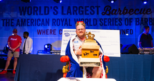 A man in a crown and cape sits on a throne, holding a trophy at a barbecue competition event.