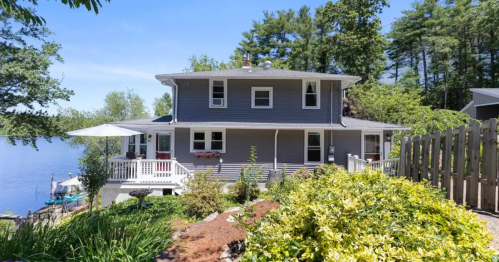 A charming gray house by a lake, surrounded by greenery and a patio with an umbrella on a sunny day.