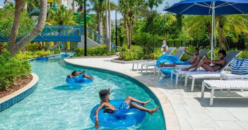 A sunny pool area with people relaxing on blue inner tubes and lounge chairs surrounded by palm trees and greenery.