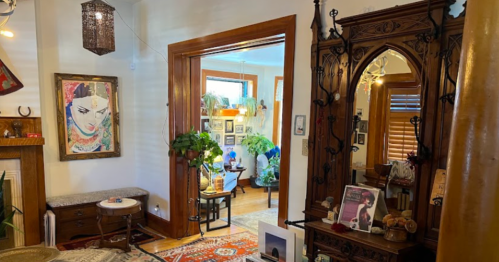A cozy, eclectic room with plants, artwork, and vintage decor, featuring a mirror and a warm, inviting atmosphere.