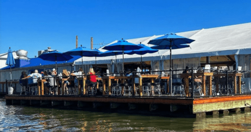 A waterfront restaurant with blue umbrellas, outdoor seating, and a large white tent under a clear blue sky.