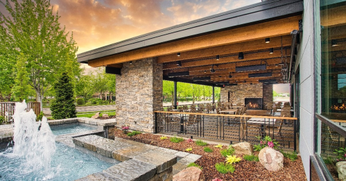 Outdoor dining area with stone accents, a fountain, and lush greenery under a colorful sunset sky.