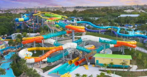 Aerial view of a colorful water park with multiple slides and pools surrounded by palm trees and greenery.