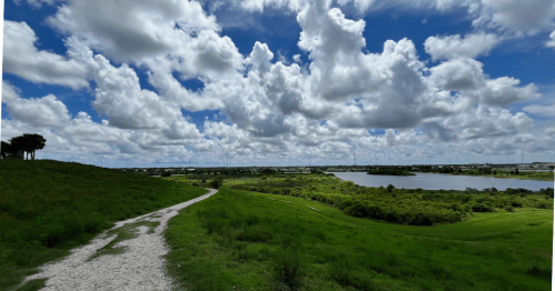 A scenic view of a winding path through green hills, with a lake and fluffy clouds in a blue sky.
