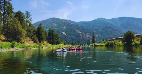 A serene river scene with people in inflatable boats surrounded by lush greenery and mountains under a clear blue sky.