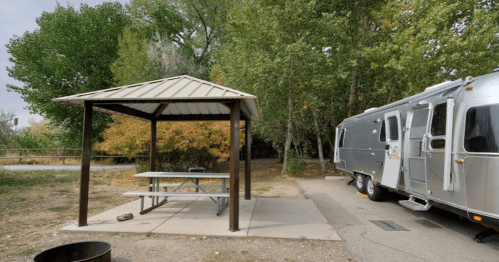 A silver Airstream trailer parked next to a shaded picnic area with a table and trees in the background.