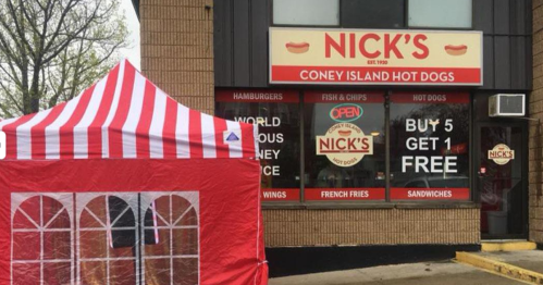 A red and white striped tent in front of Nick's Coney Island Hot Dogs, featuring a menu and promotional signs.