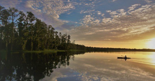 A serene lake at sunset, with a lone kayaker paddling amidst trees and colorful clouds reflecting on the water.