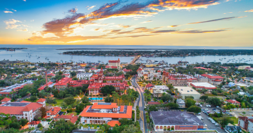 Aerial view of a coastal town at sunset, featuring colorful buildings, a marina, and a vibrant sky.