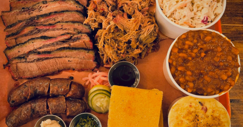 A platter of barbecue featuring sliced brisket, pulled pork, sausage, coleslaw, baked beans, cornbread, and pickles.