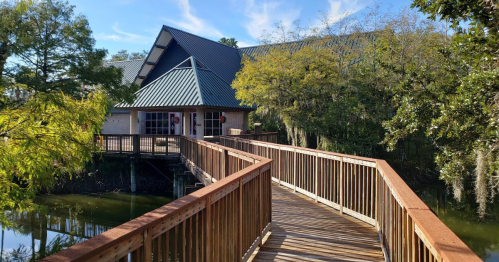 A wooden walkway leads to a building surrounded by trees and water, under a clear blue sky.