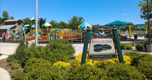 A community park sign in front of a colorful playground with green trees and blue skies in the background.