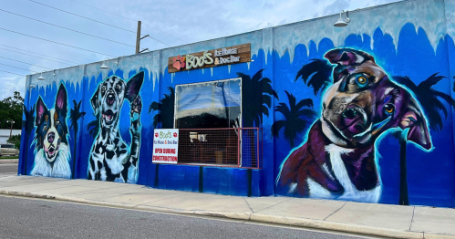 Colorful mural of three dogs on a blue wall, featuring palm trees and a sign for a pet-friendly business.
