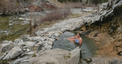 A person relaxes in a natural hot spring by a river, surrounded by rocky terrain and sparse vegetation.