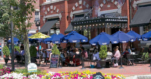 Outdoor dining scene at a restaurant with blue umbrellas, surrounded by flowers and people enjoying their meals.