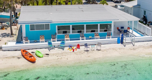 A beach house with colorful chairs and kayaks on the sandy shore, surrounded by clear turquoise water.