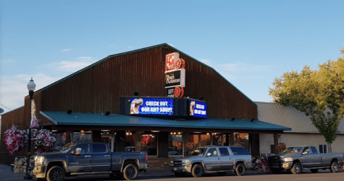 A rustic restaurant building with a green roof, featuring bright signs and parked trucks in front.
