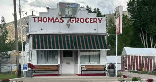A quaint grocery store named "Thomas Grocery" with a striped awning and benches outside, surrounded by trees.