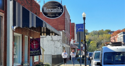 A sunny street scene featuring shops, including Willow Spring Mercantile, with two people walking and a parked vehicle.