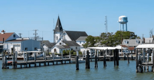 A peaceful waterfront scene featuring a church, water tower, and boats docked at a marina under a clear blue sky.